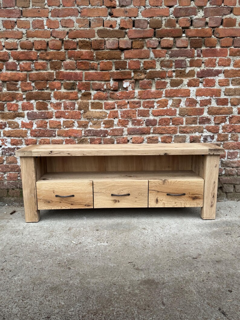 May include: A light-colored wooden media console with three drawers and a shelf. The console has a rectangular shape and is made of natural wood. The drawers have black handles. The background is a brick wall.