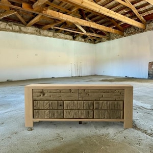 May include: A wooden dresser with a light-colored frame and a textured, darker wood front. The dresser has two rows of drawers, each with a unique, rustic design. The piece is set in a room with a concrete floor and exposed wooden beams.