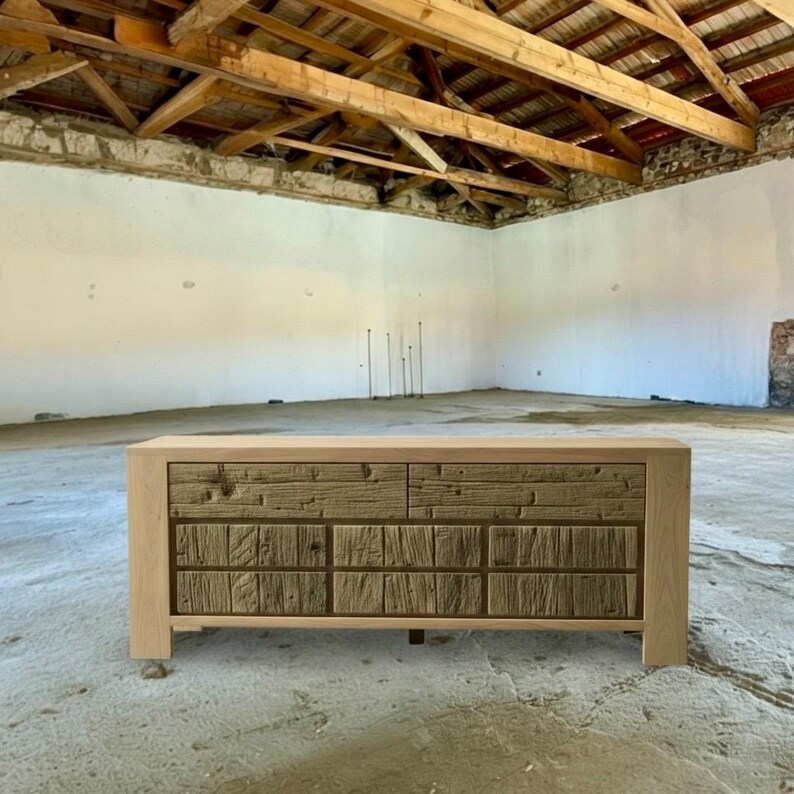 May include: A wooden dresser with a light-colored frame and a textured, darker wood front. The dresser has two rows of drawers, each with a unique, rustic design. The piece is set in a room with a concrete floor and exposed wooden beams.