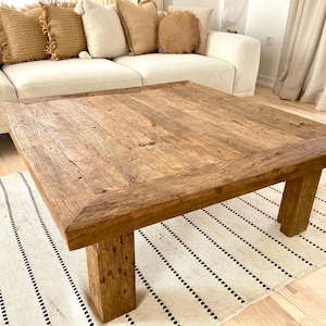 May include: A square, rustic wooden coffee table with a natural finish. The table has a thick, textured top and sturdy, block-style legs. It sits on a cream-colored rug with black stripes. The table is surrounded by a cream-colored sofa and decorative pillows.