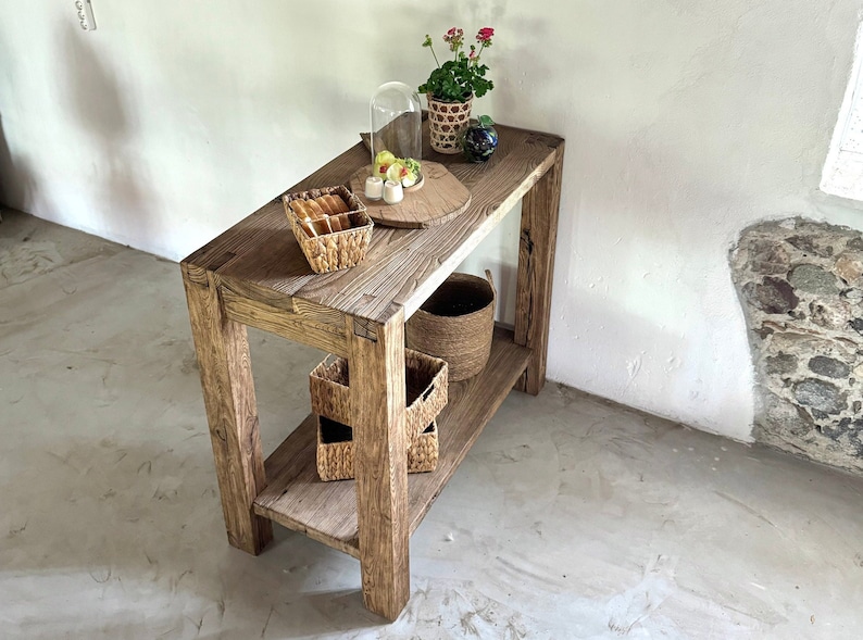 K&ouml;nnte beinhalten: Ein rustikaler Holz-Konsolentisch mit zwei Ablagen. Der Tisch ist mit einer Glasglocke, einem kleinen Teller mit Essen, einer Topfpflanze und mehreren geflochtenen K&ouml;rben dekoriert.