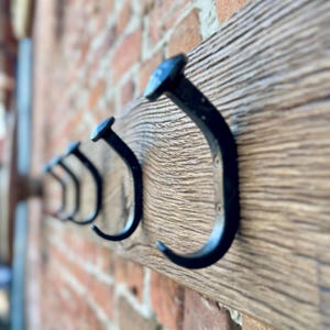 May include: A row of black metal hooks mounted on a rustic wooden board. The hooks are in the shape of a J and are attached to the board with screws. The board is made of reclaimed wood and has a natural finish.