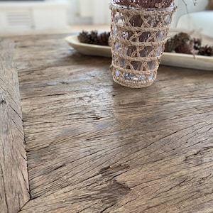 May include: A rustic, weathered wooden coffee table with a natural finish. A clear glass vase wrapped in woven twine sits on the table, along with a wooden bowl holding pinecones. The table's surface displays prominent wood grain.