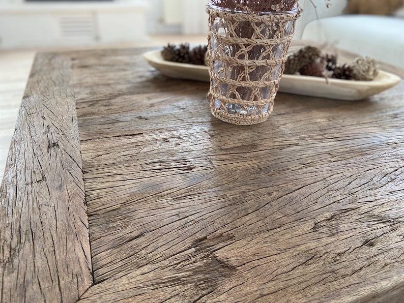 May include: A rustic, weathered wooden coffee table with a natural finish. A clear glass vase wrapped in woven twine sits on the table, along with a wooden bowl holding pinecones. The table's surface displays prominent wood grain.