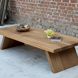 Puede incluir: Mesa de centro rectangular de madera clara con patas anguladas. La mesa tiene un acabado natural. Un cuenco de madera con objetos decorativos, un peque&ntilde;o jarr&oacute;n con flores amarillas y un libro est&aacute;n sobre la mesa. La mesa mide unos 150 cm de largo.