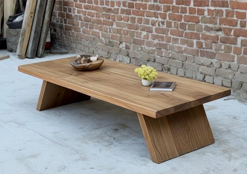 Puede incluir: Mesa de centro rectangular de madera clara con patas anguladas. La mesa tiene un acabado natural. Un cuenco de madera con objetos decorativos, un peque&ntilde;o jarr&oacute;n con flores amarillas y un libro est&aacute;n sobre la mesa. La mesa mide unos 150 cm de largo.