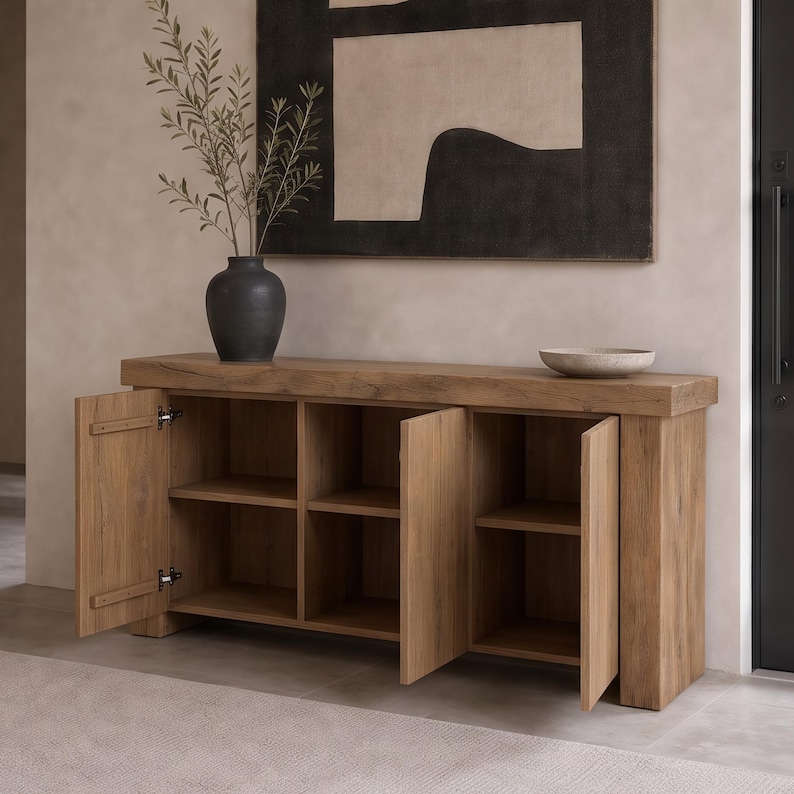 Peut inclure: Un buffet rectangulaire en bois avec une finition grain de bois naturel, dot&eacute; de quatre portes d'armoire ouvertes r&eacute;v&eacute;lant des &eacute;tag&egrave;res int&eacute;rieures. Un vase gris fonc&eacute; avec une branche feuillue et un bol en pierre peu profond sont pos&eacute;s dessus.