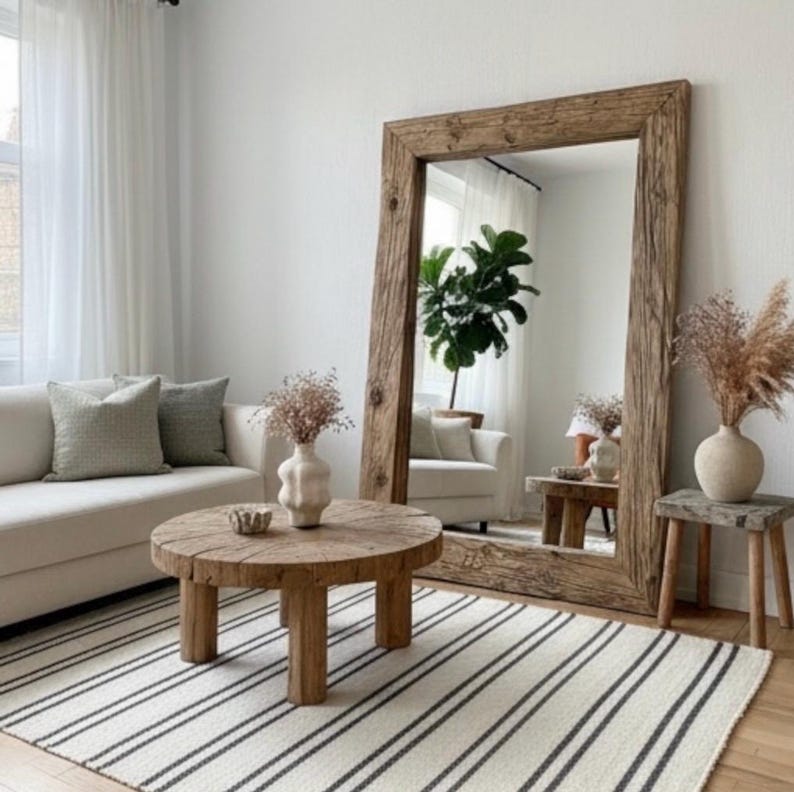 May include: A living room with a large wooden-framed mirror, a round wooden coffee table, and a striped rug. A white sofa with green cushions and vases with dried flowers are also present, creating a cozy atmosphere.