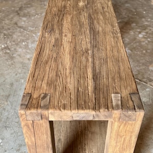 Peut inclure: Banc en bois rectangulaire avec une finition naturelle et patin&eacute;e. Le banc pr&eacute;sente un grain de bois visible et est construit avec des assemblages &agrave; tenons et mortaises. Les pieds sont robustes et le design g&eacute;n&eacute;ral est rustique et minimaliste.