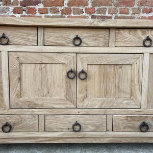 May include: A light-colored wooden cabinet with multiple drawers and doors. The cabinet has dark metal ring pulls on the drawers and doors. The wood grain is visible, and the cabinet is set against a brick wall.