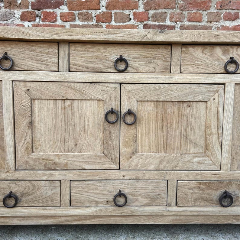 May include: A light-colored wooden cabinet with multiple drawers and doors. The cabinet has dark metal ring pulls on the drawers and doors. The wood grain is visible, and the cabinet is set against a brick wall.