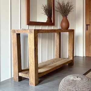 May include: A rustic wooden console table with a lower shelf. A terracotta vase with dried plants sits on top. A wooden framed mirror hangs on the wall behind the table. The room has a neutral color palette.