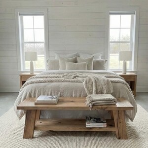 May include: A neutral-toned bedroom with a bed, bench, and rug. The bed has a beige comforter and multiple pillows. A wooden bench sits at the foot of the bed, holding books and a folded blanket. Two bedside tables with lamps flank the bed.