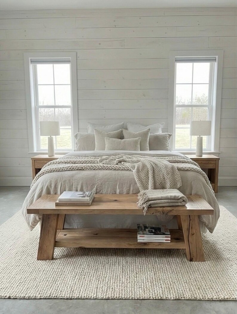 May include: A neutral-toned bedroom with a bed, bench, and rug. The bed has a beige comforter and multiple pillows. A wooden bench sits at the foot of the bed, holding books and a folded blanket. Two bedside tables with lamps flank the bed.