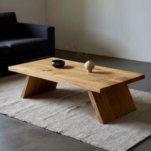 Puede incluir: Mesa de centro rectangular de madera clara con patas anguladas. Un peque&ntilde;o cuenco negro y un jarr&oacute;n blanco con una ramita de flores secas est&aacute;n sobre la mesa. La mesa descansa sobre una alfombra gris claro.
