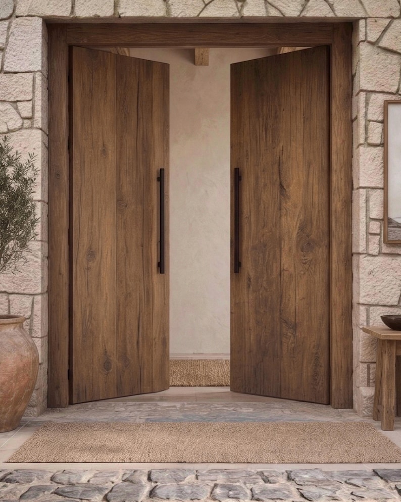 May include: A pair of tall, double wooden doors with a natural brown wood grain finish and long, vertical black metal handles. The doors are set within a light-colored stone archway, featuring a textured surface and a visible wood plank construction.