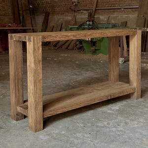 Peut inclure: Une console en bois fabriqu&eacute;e &agrave; la main avec une finition naturelle. La table pr&eacute;sente un plateau rectangulaire et une &eacute;tag&egrave;re inf&eacute;rieure, soutenus par quatre pieds robustes. Le grain du bois est visible, mettant en valeur la beaut&eacute; naturelle du mat&eacute;riau. La table mesure environ 122 cm de long.