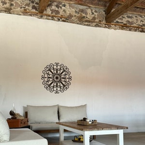 May include: A white coffee table with a wooden top and a lower shelf. The table is in a living room with a white sofa and a decorative metal wall hanging.