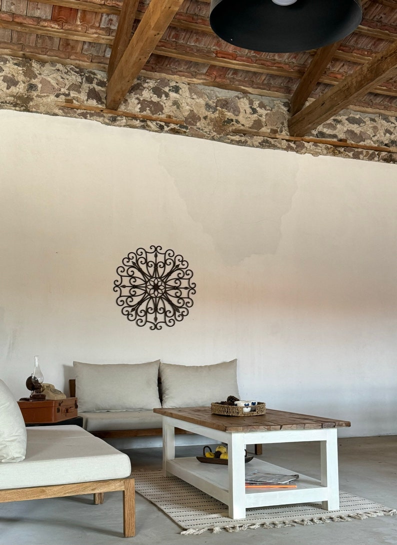 May include: A white coffee table with a wooden top and a lower shelf. The table is in a living room with a white sofa and a decorative metal wall hanging.