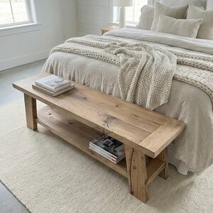 May include: A rustic wooden bench with a lower shelf, placed at the foot of a bed. The bench is made of light-colored wood and has a natural finish. Two stacks of books and magazines are on the bench.