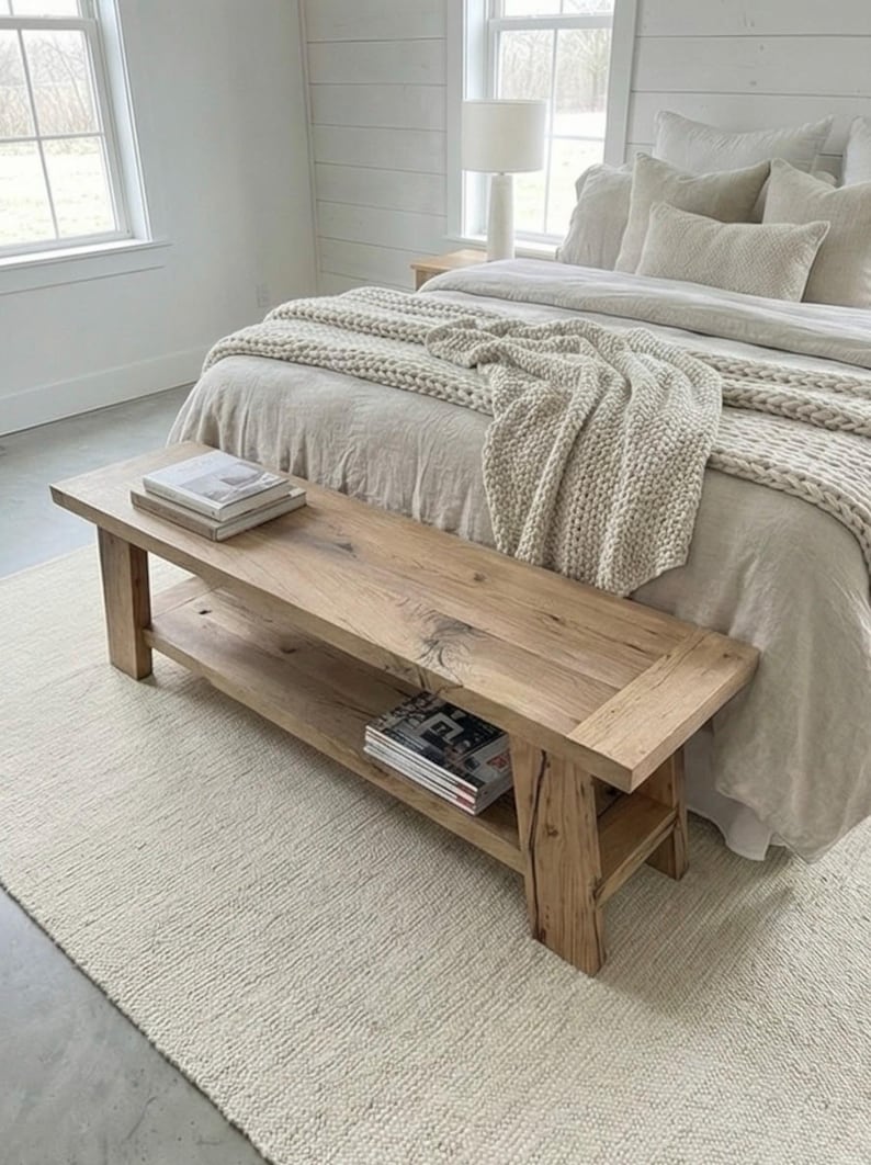 May include: A rustic wooden bench with a lower shelf, placed at the foot of a bed. The bench is made of light-colored wood and has a natural finish. Two stacks of books and magazines are on the bench.