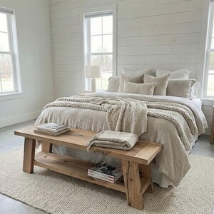 May include: A bedroom with a neutral color palette. A wooden bench sits at the foot of a bed with beige bedding and a chunky knit throw. A woven rug and white paneled walls complete the cozy, inviting space.