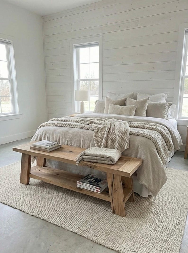 May include: A bedroom with a neutral color palette. A wooden bench sits at the foot of a bed with beige bedding and a chunky knit throw. A woven rug and white paneled walls complete the cozy, inviting space.