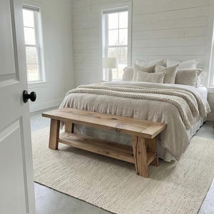May include: A bedroom with a neutral color palette. The bed is covered with beige bedding and pillows, and a wooden bench sits at the foot of the bed. A cream-colored rug covers the floor, and the walls are white.
