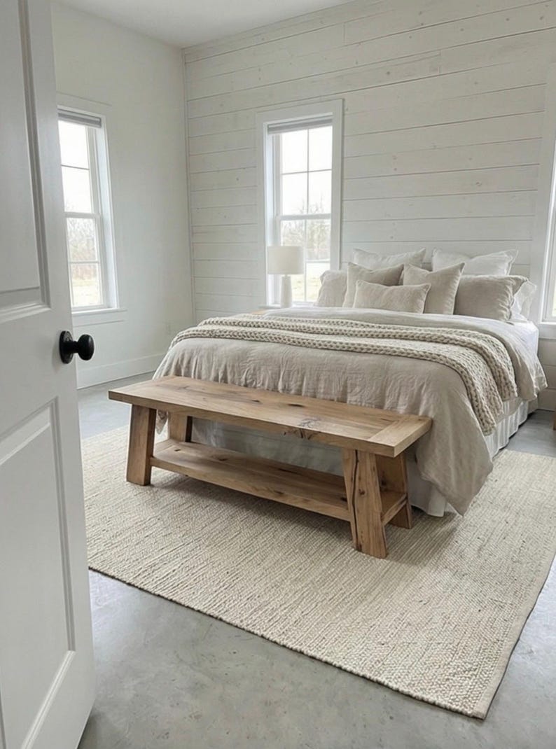 May include: A bedroom with a neutral color palette. The bed is covered with beige bedding and pillows, and a wooden bench sits at the foot of the bed. A cream-colored rug covers the floor, and the walls are white.