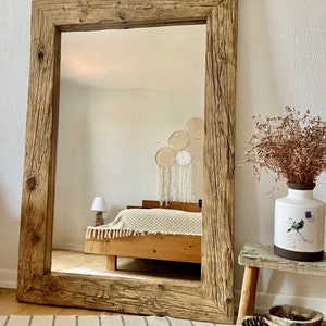May include: A large rectangular mirror with a distressed wooden frame. The frame is made from reclaimed wood, showing the natural grain and knots. The mirror reflects a bedroom scene, with a bed and decorative items, creating a warm and inviting atmosphere.