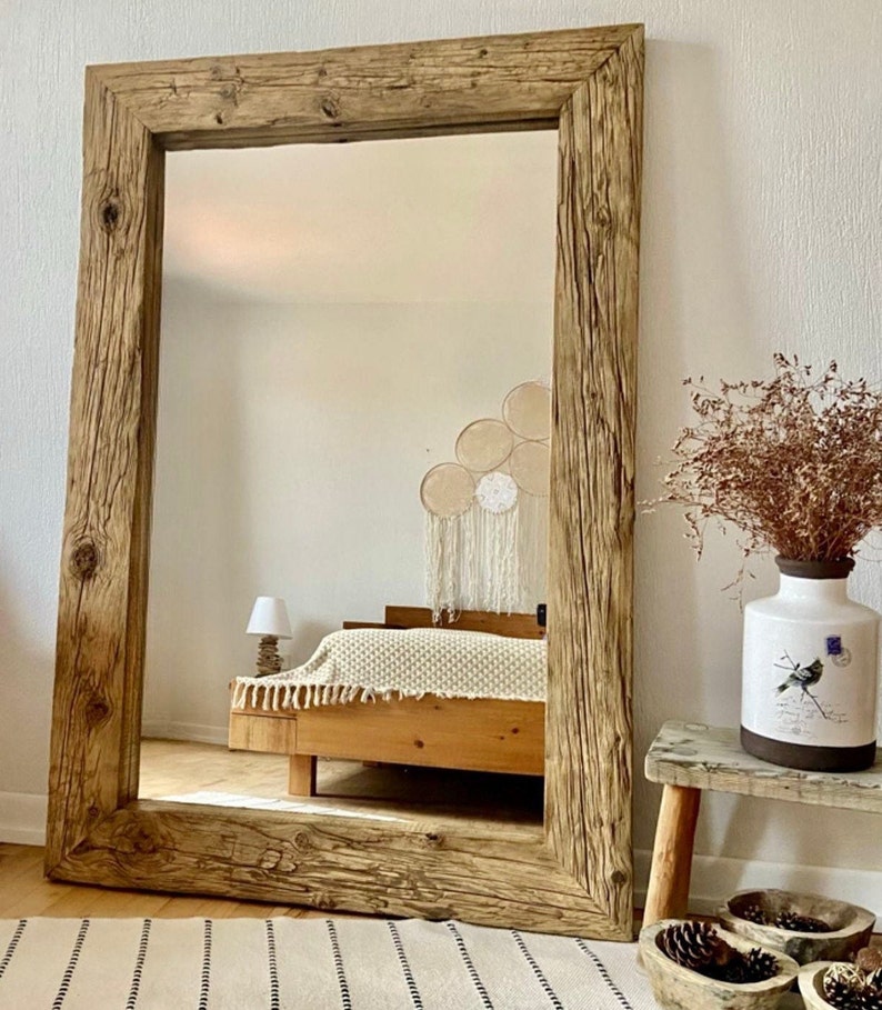 May include: A large rectangular mirror with a distressed wooden frame. The frame is made from reclaimed wood, showing the natural grain and knots. The mirror reflects a bedroom scene, with a bed and decorative items, creating a warm and inviting atmosphere.