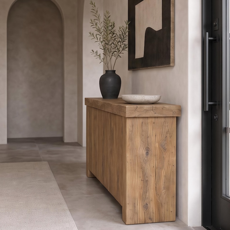 Peut inclure: Une console rectangulaire en bois au grain naturel, surmont&eacute;e d'un vase noir mat contenant des branches feuillues vertes et d'un bol en pierre peu profond &agrave; la texture granuleuse, le tout dispos&eacute; contre un mur aux tons neutres.