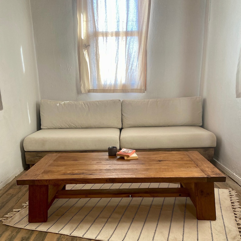 May include: A living room setting with a wooden coffee table and a cream-coloured sofa. The coffee table has a natural finish. A small rug with black stripes is on the floor. A book and a cup are on the table. The room has a window with a curtain.