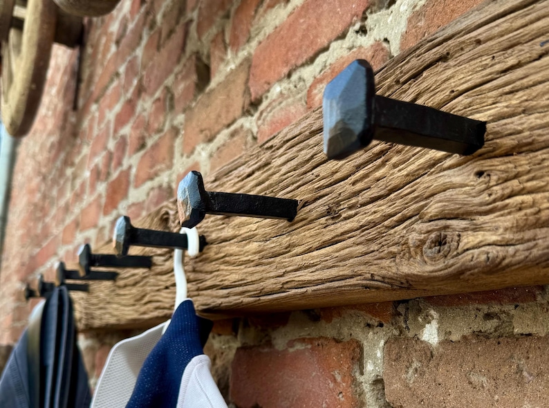 May include: A rustic wooden coat rack with five black metal hooks. The hooks are shaped like a diamond and are hammered into the wood. The rack is mounted on a brick wall.