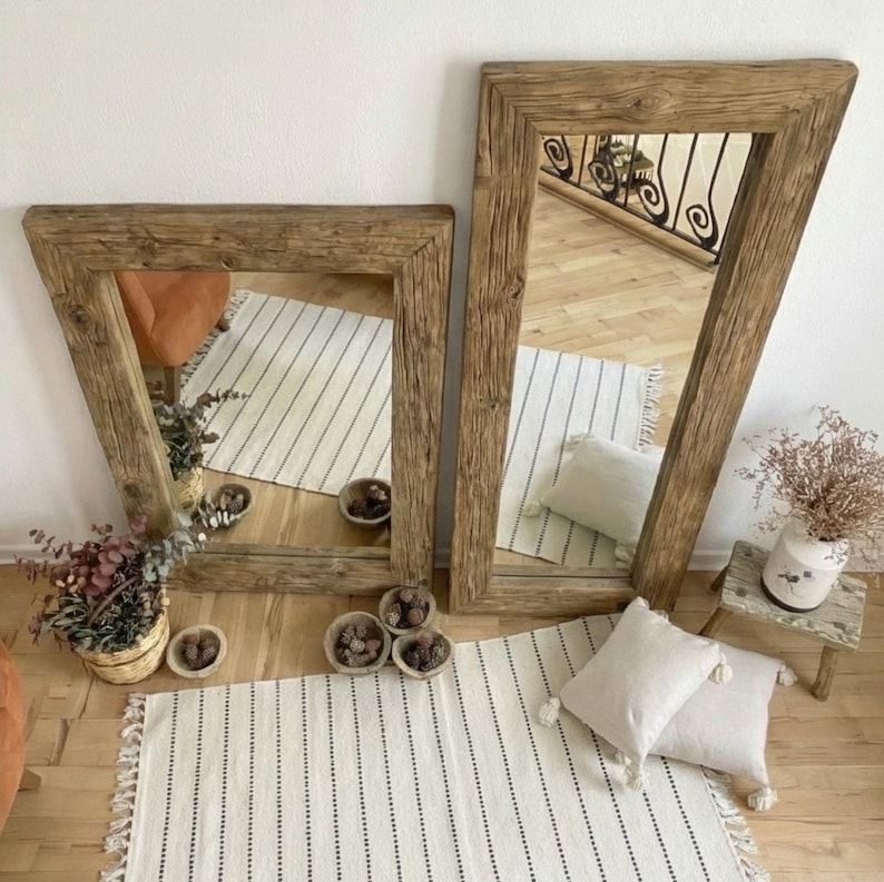 May include: Two wooden-framed mirrors with a natural, weathered finish. The mirrors reflect a room with a striped rug, pillows, and decorative items. The frames have a thick, textured appearance, adding a touch of farmhouse style.