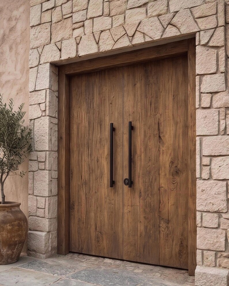 May include: A set of double doors made of brown wood with a visible grain pattern, featuring two long, vertical black metal handles. The doors are framed by light-colored, irregular stone blocks that form an archway above the entrance.