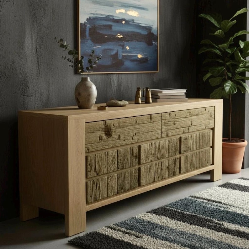 May include: A light oak wood sideboard with six drawers, featuring a textured, rustic wood panel design on the drawer fronts. The sideboard is in a room with a painting, a plant, and a striped rug.