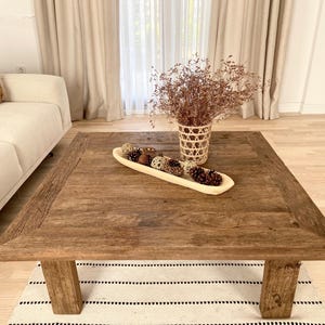 May include: A rustic, square wooden coffee table with a distressed finish. The table features thick legs and a decorative wooden bowl filled with pine cones and decorative spheres. A woven basket with dried flowers sits on the table.