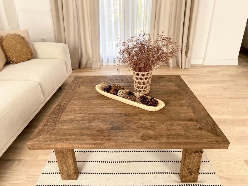 May include: A rustic, square wooden coffee table with a distressed finish. The table features thick legs and a decorative wooden bowl filled with pine cones and decorative spheres. A woven basket with dried flowers sits on the table.