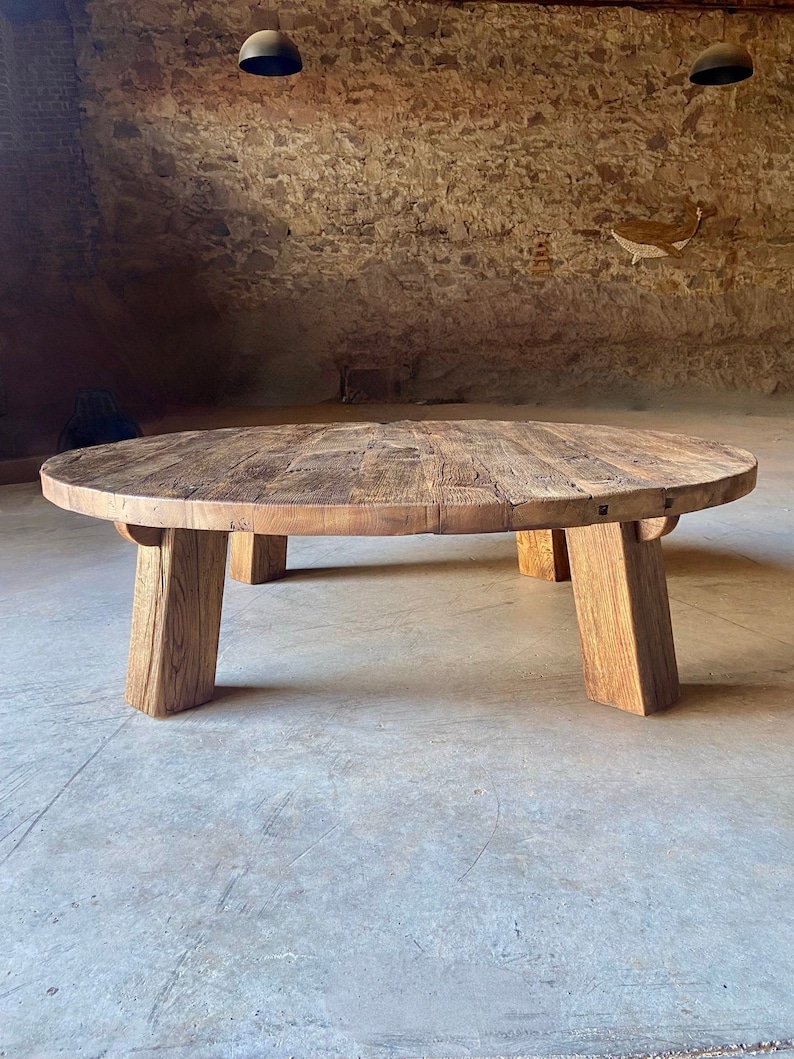 May include: A large, round wooden coffee table with a natural wood finish and four sturdy legs. The table is made of reclaimed wood and has a rustic, farmhouse style.
