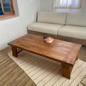 May include: A rectangular, wooden coffee table with a natural finish. The table is set on a cream-coloured rug with black stripes. A small book and a dark-coloured cup sit on the table. The table is in front of a beige sofa.