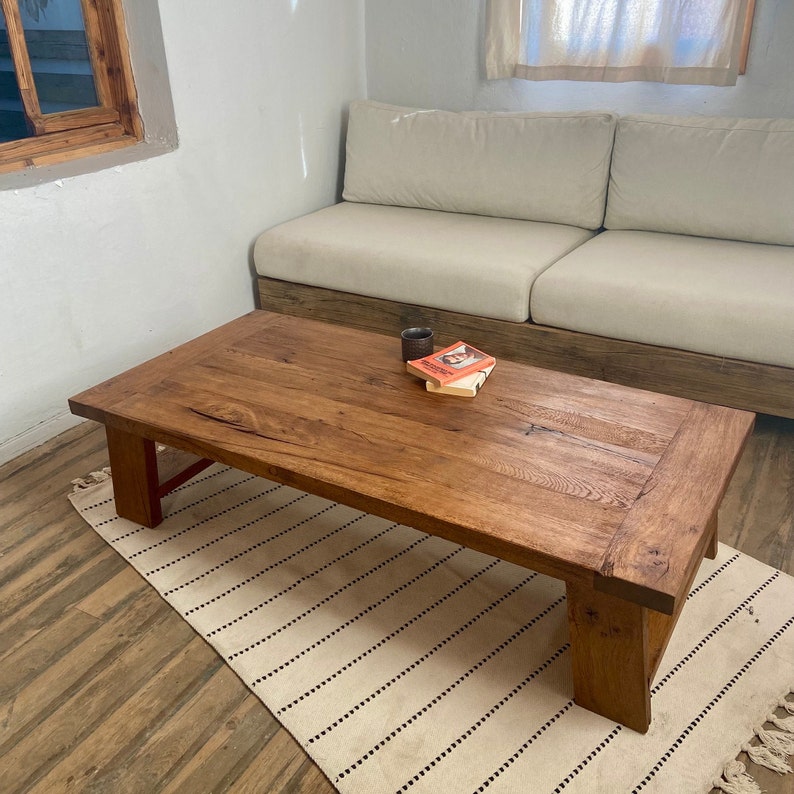 May include: A rectangular, wooden coffee table with a natural finish. The table is set on a cream-coloured rug with black stripes. A small book and a dark-coloured cup sit on the table. The table is in front of a beige sofa.