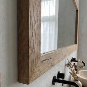 May include: A rectangular mirror with a natural wood frame. The frame has a rustic, textured appearance with visible wood grain. The mirror is mounted on a light-colored wall, reflecting a window with sheer curtains. Black faucets are visible.