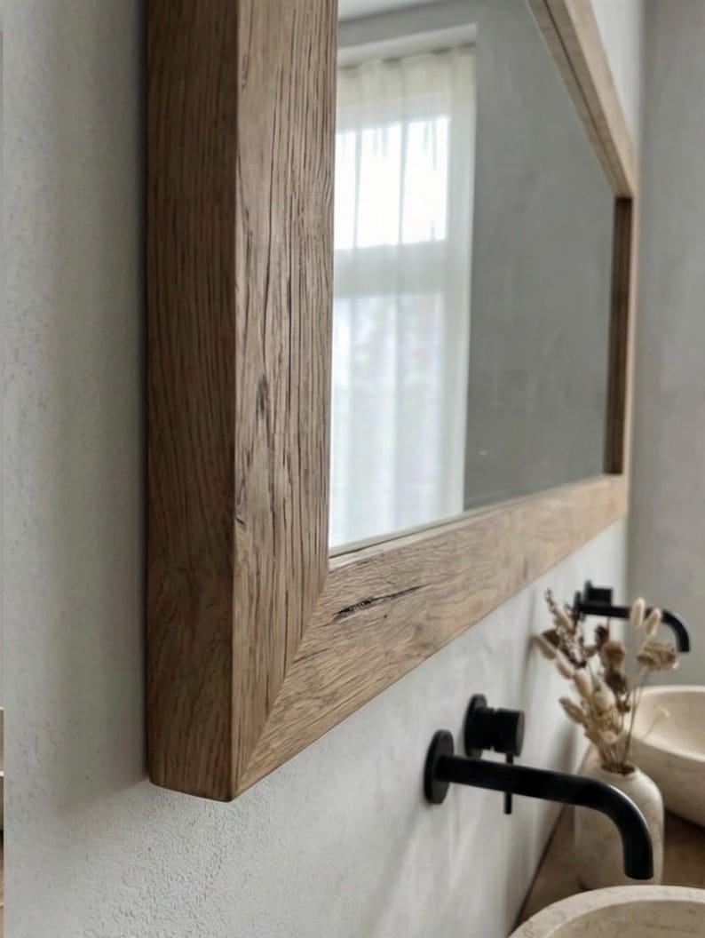 May include: A rectangular mirror with a natural wood frame. The frame has a rustic, textured appearance with visible wood grain. The mirror is mounted on a light-colored wall, reflecting a window with sheer curtains. Black faucets are visible.