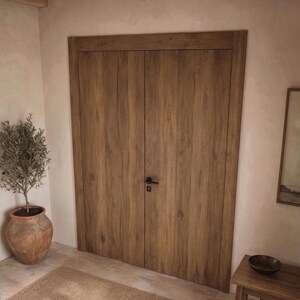 May include: A set of double doors featuring a medium brown wood grain finish with vertical planks and a matte black metal handle. The doors are framed by a matching wood trim and are set within a light-colored wall with a smooth texture.