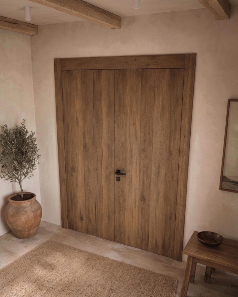 May include: A set of double doors featuring a medium brown wood grain finish with vertical planks and a matte black metal handle. The doors are framed by a matching wood trim and are set within a light-colored wall with a smooth texture.