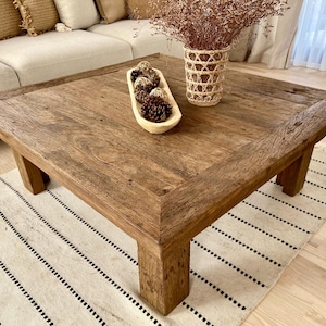 May include: A large, square, rustic wooden coffee table with a distressed finish. The table is adorned with a decorative wooden bowl filled with pinecones and a woven vase with dried flowers. The table sits on a striped rug.