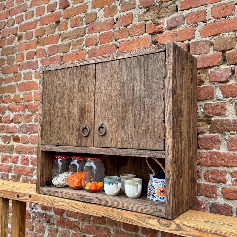 Puede incluir: Un armario de pared de madera r&uacute;stica con dos puertas y un estante. El armario tiene dos tiradores de anillo de metal y est&aacute; pintado de un color marr&oacute;n oscuro. El estante est&aacute; lleno de tarros de vidrio y tazas.