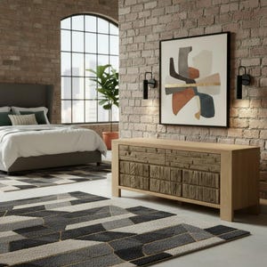 May include: A bedroom scene featuring a bed with a gray headboard, white bedding, and a geometric patterned rug. A wooden dresser with textured drawer fronts and an abstract art piece hang on the brick wall.