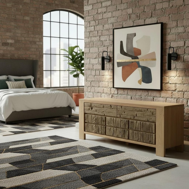 May include: A bedroom scene featuring a bed with a gray headboard, white bedding, and a geometric patterned rug. A wooden dresser with textured drawer fronts and an abstract art piece hang on the brick wall.